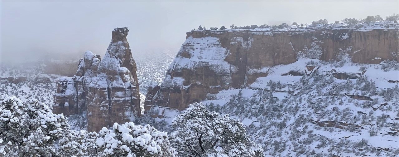 Snow-covered red-rock spires and cliffs stand among white mist and snow-laden trees.