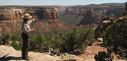 Standing towers of red-orange sandstone intersperse a green blanket of shrubs and trees. A ranger in uniform directs attention with open hands.