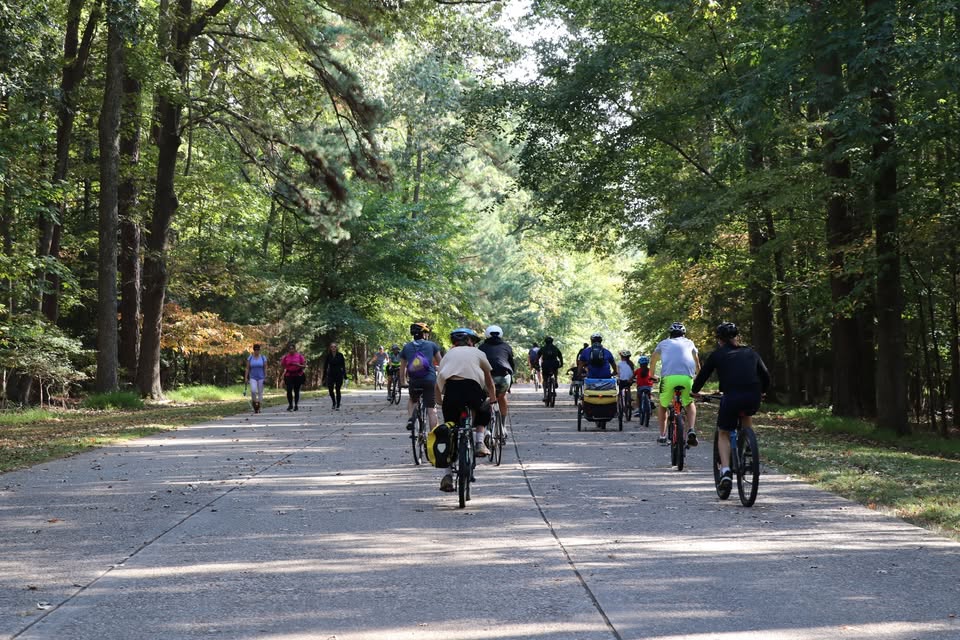 Approximately fifteen bicycle riders and three pedestrians are on a segment of the Colonial Parkway