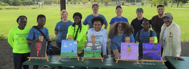 12 people smiling and posing behind five landscape paintings.