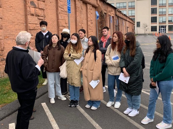 A group of 10 students watching a listening to a woman showing them a piece of paper.