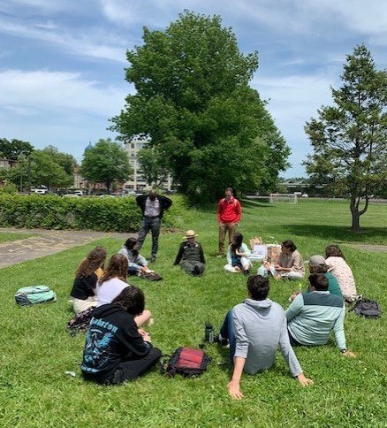 A group of students sitting in a circle outside on grass listening to a ranger sitting with them.