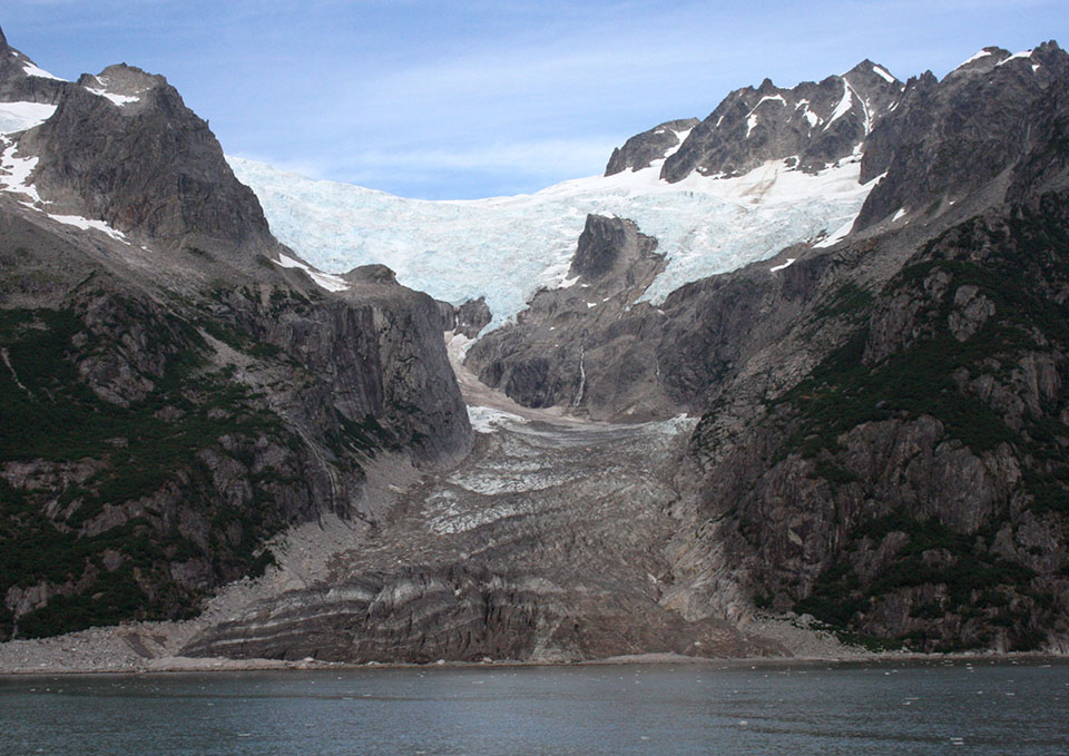 The bottom fifthof the picture is water.  A blue colored glacier flows over a mountain at the top of the picture, and down to the water.  The glacier is narrow in the middle of the image as it flows between two rocky outcroppings.