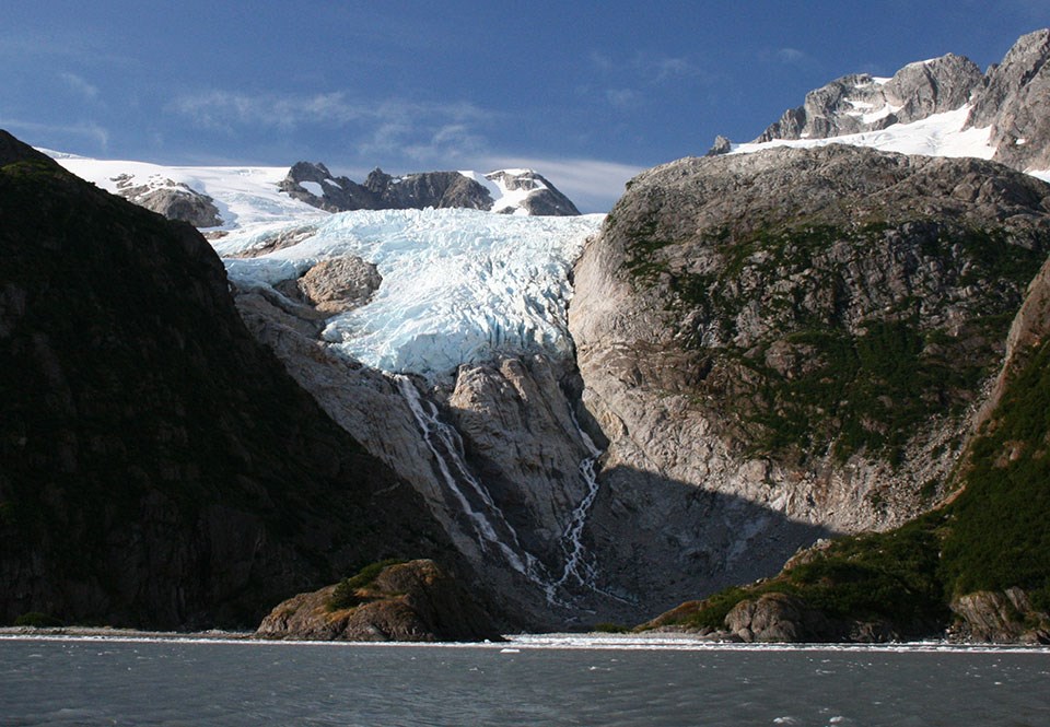 A blue colored glacier flows over a mountainside and down to the water.  There are rocks and land in the water in front of the glacier