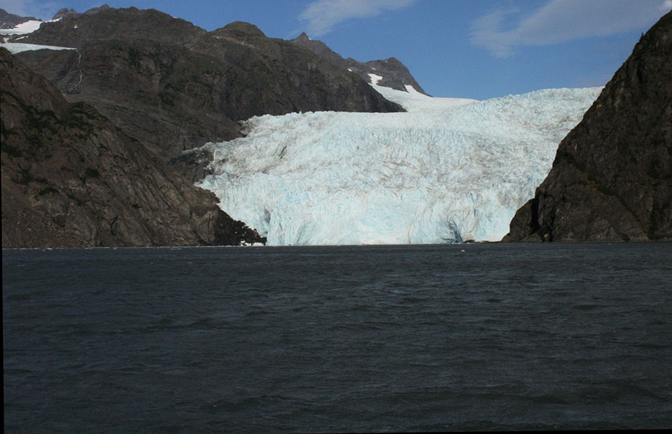 A black and white photo.  The bottom half of the picture is water.  There are mountainsides on the right and left of the picture.  In the middle is a large glacier that meets the water.