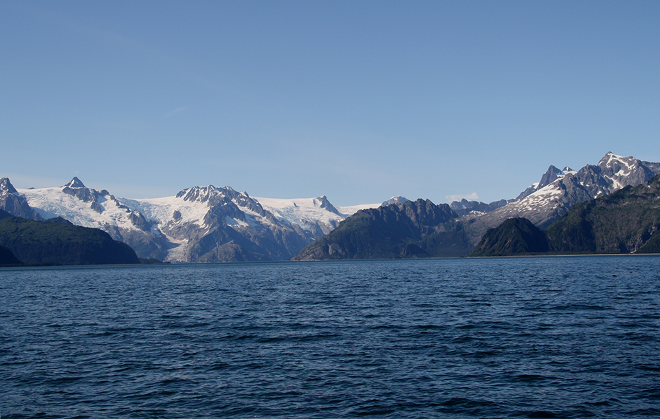 A black and white photo.  The bottom half of the picture is water. In the center of the image is a glacier.  On the left and right of hte glacier are rocky outcroppings.  In the background are snow covered mountains