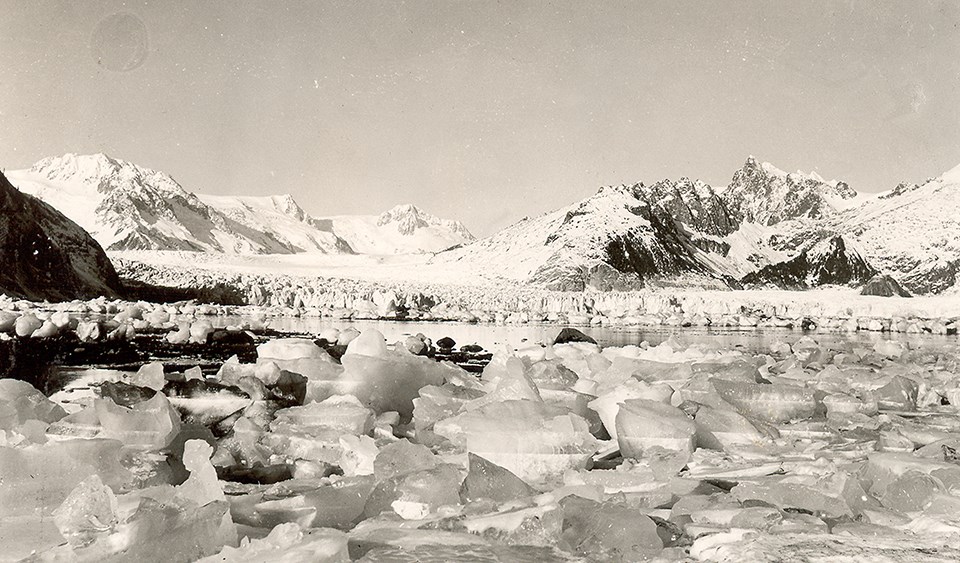 A black and white photo.  Chunks of ice are in the foreground of the image.  BEhind them is a thin strip of water, and then the face of a glacier.  There are snow covered mountains on behind the glacier