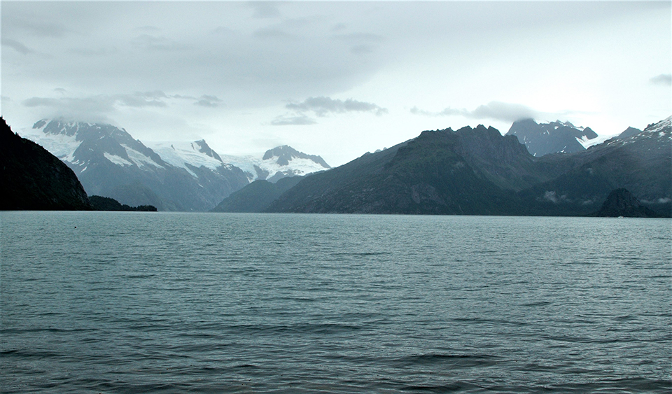 A black and white photo.  Chunks of ice are in the foreground of the image.  BEhind them is a thin strip of water, and then the face of a glacier.  There are snow covered mountains on behind the glacier