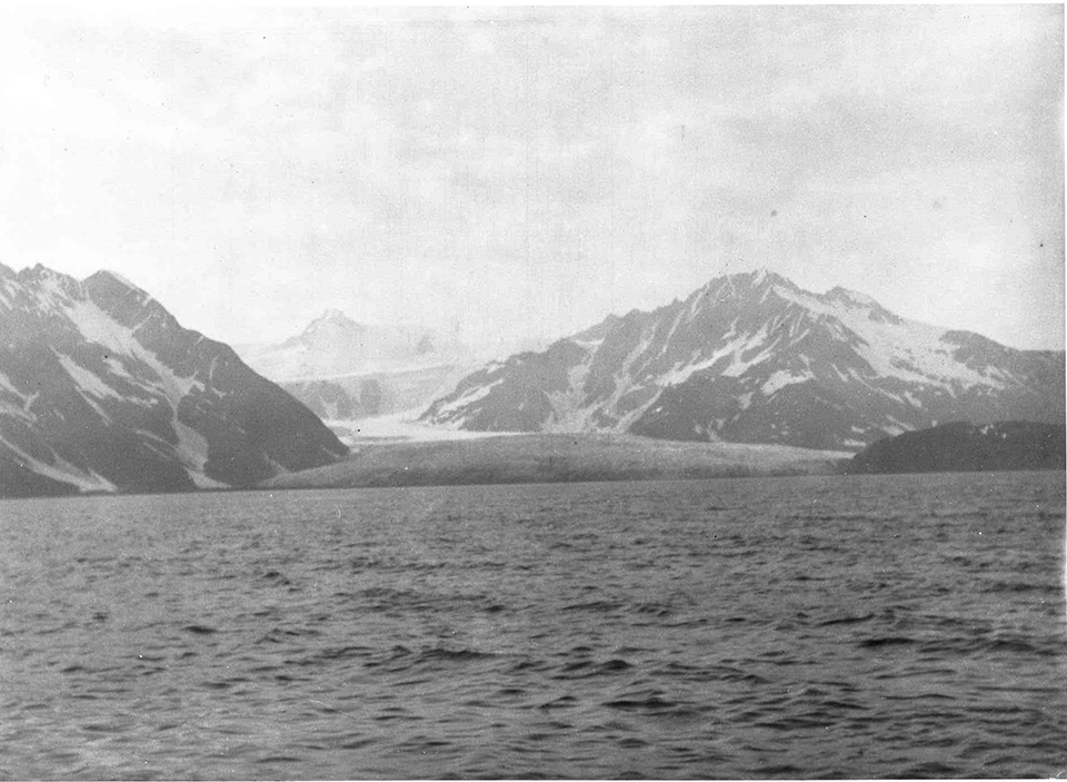 A black and white photo. The bottom half of the picture is water.  Two mountains are on either side of htei mage, and a third mountain is faint in the background.  A white glacier is in the center of the image and comes to the waters edge