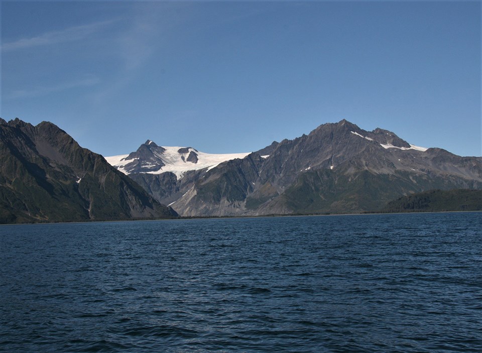A black and white photo. The bottom half of the picture is water.  Two mountains are on either side of htei mage, and a third mountain is faint in the background.  A white glacier is in the center of the image and comes to the waters edge