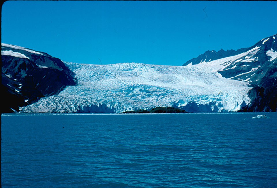 A tidewater in 1990. The glacier is in the center of the image.  The bottom of the glacier is at the edge of water.  the glacier is between two mountainsides