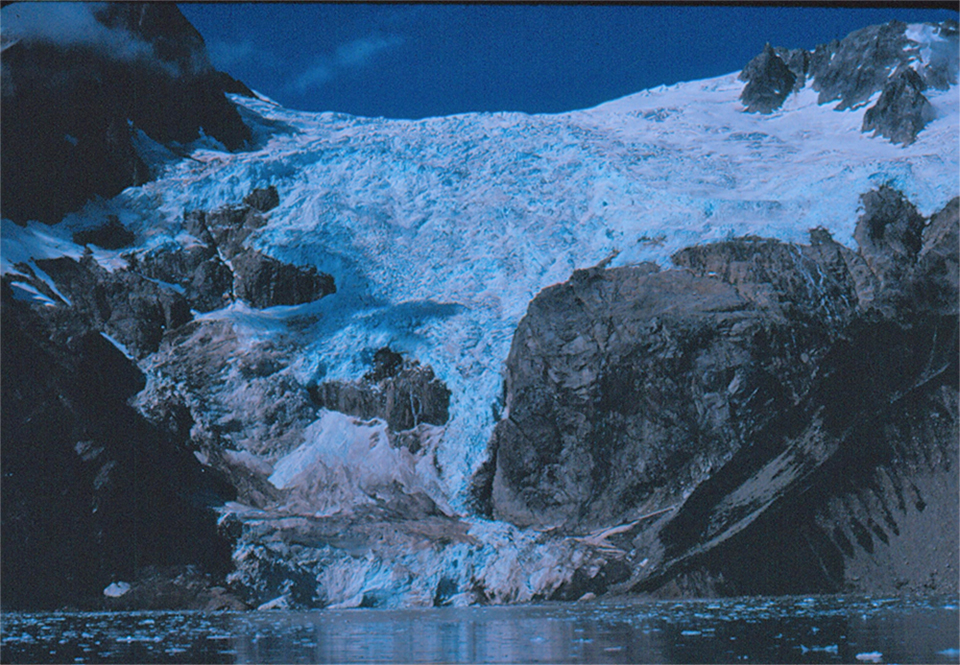 A blue colored glacier flows over a mountainside.  Center right is a large rocky outcropping from undre thglacier.