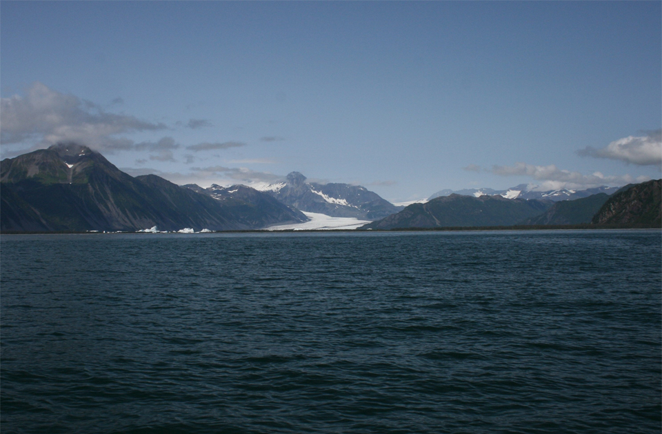 A black and white photo.  The bottom half of the picture is water.  In the top half there is a large glacier in the center of center of the picture.  There are mountains on the right edge and left edge of the picture to the sides of the glacier.