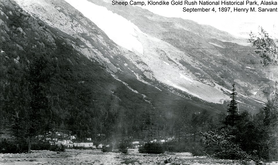 Black and white image of tents and buildings at the base of a mountain with a glacier above it