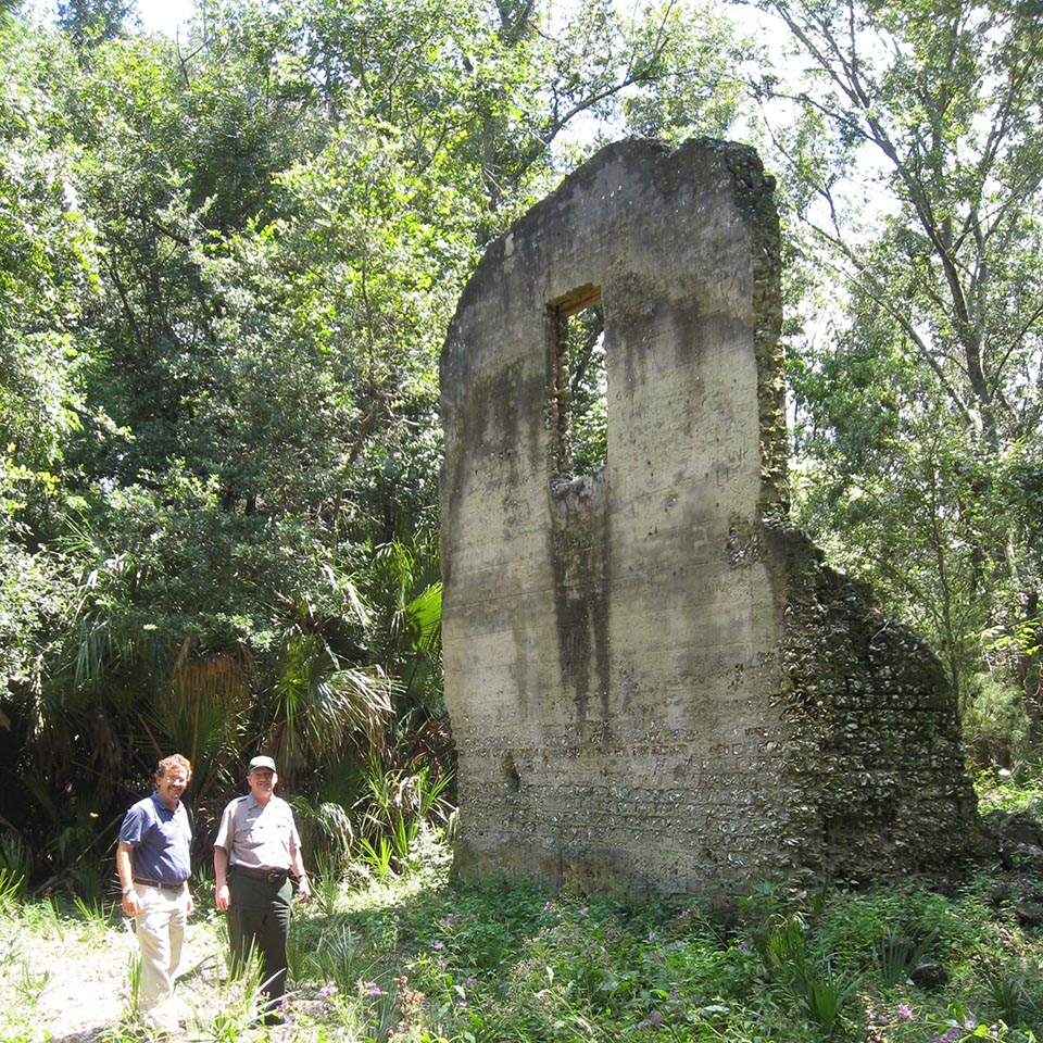 two story tabby wall in the woods with a second story window hole and worn walls on the first floor section, two men stand to the side