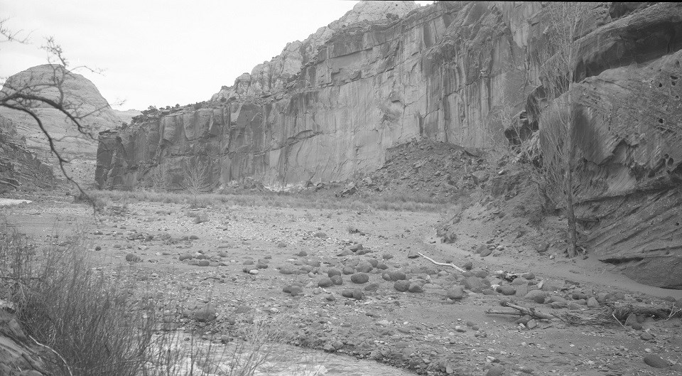 Black and white photo of valley below large rock cliffs, with a river to the lower left.