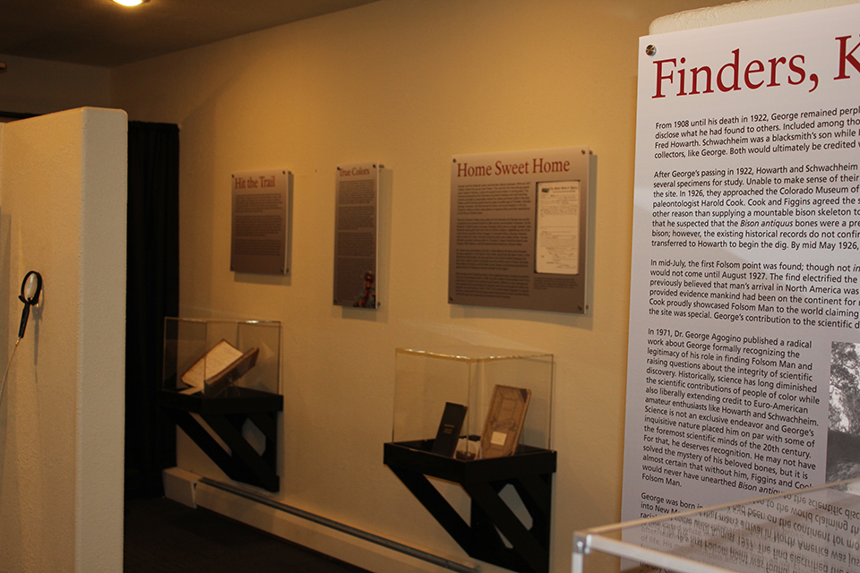 A television sits inside a wall of wood paneling.  On both sides are speakers also embedded in the wall.  Five chair backs face the camera.  To the left is an exit illuminated by daylight.