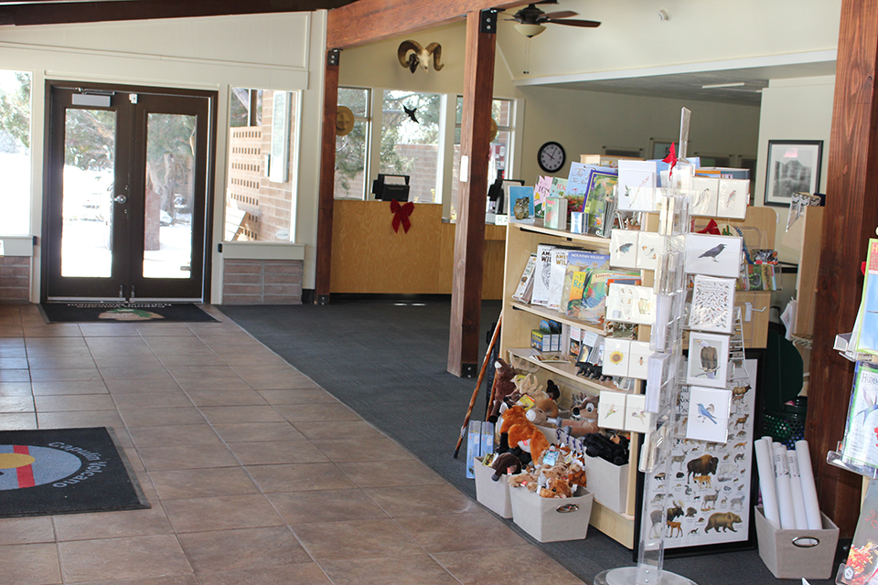 A visitor center foyer is dappled in sunlight.  Along the righ the American flag stands posted at the entry to the main building.  Ahead two benches line each side of the walkway.  To the left a brick wall of windows faces outside.