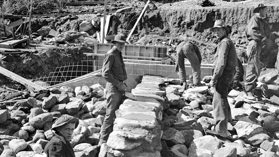 Black and white photo of six men stacking rocks near a river bank.