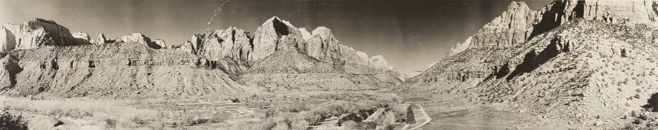 Panoramic black and white photo looking up Zion Canyon, structures are seen near the river.
