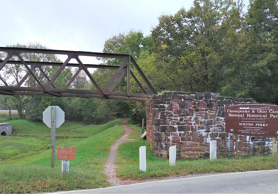 A photo of a dirt path surrounded by grass running beneath a metal bridge.