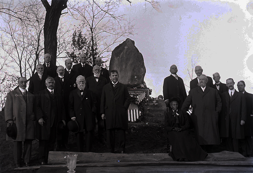 A group of men in dress coats and a seated woman in front of a stone marker
