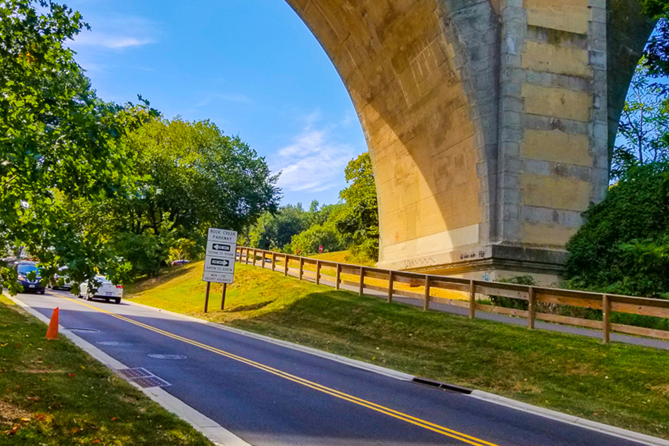 Road with potholes under bridge with footpath nearby.