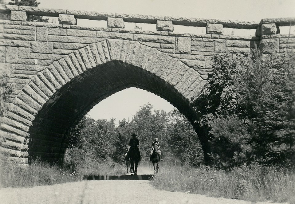 Two horseback riders under a stone bridge