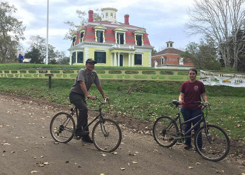 A man and a woman sit on bicycles in front of a bright yellow house on a hill.