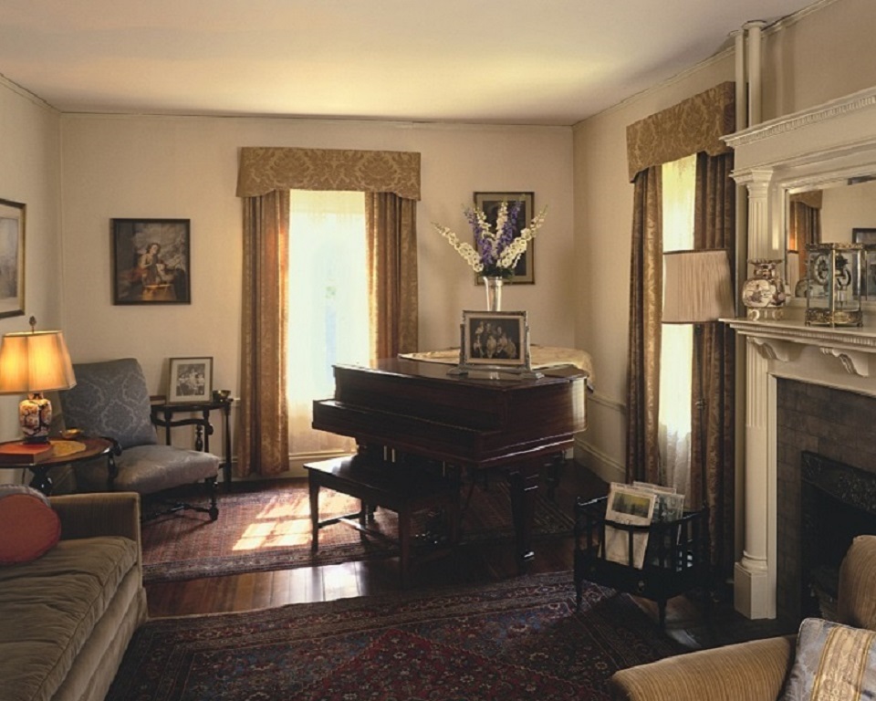 Floral curtains hang over two windows in the back of the room.  A TV set sits between the windows.  A gas fireplace is to the right.  A portrait of JFK is on the left back wall.  A chair and couch is to the left.