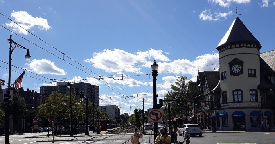 A view of Coolidge Corner at the intersection of Harvard and Beacon Streets.  The SS Pierce Building with clock tower is on the left.  A streetcar system runs alongside the SS Pierce to the left.  People are boarding the streetcar.