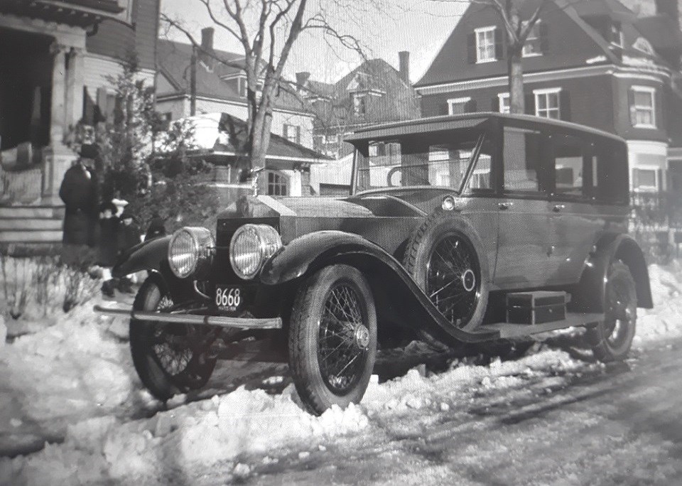 A black and white photo.  A large car sits in the forefront of the photo.  In the back left corner is a set of steps.  On the steps are a man in dark clothing with three small children.