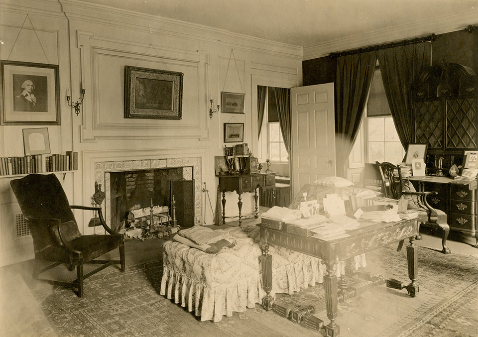 Black and white image of room with table piled with books and chaise at center.