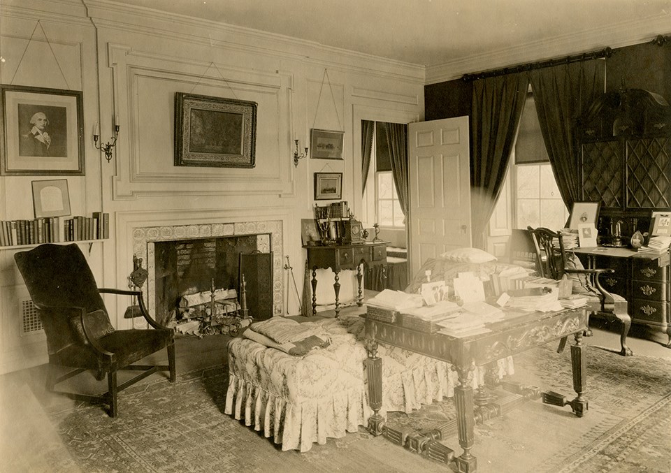 Black and white image of room with table piled with books and chaise at center.
