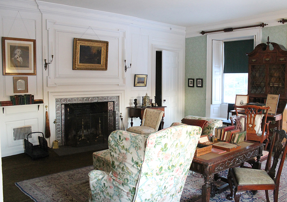 Black and white image of room with table piled with books and chaise at center.