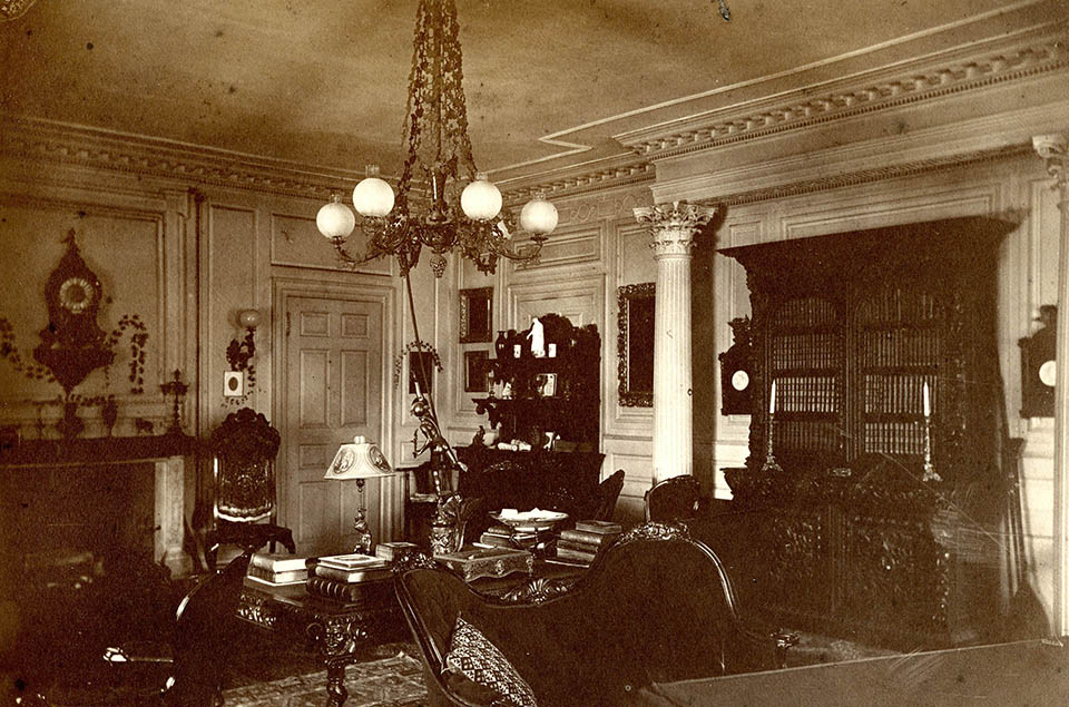 Black and white photograph of Victorian interior with ornate furniture including bookcase and table piled with books.