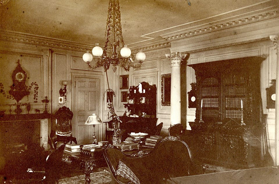 Black and white photograph of Victorian interior with ornate furniture including bookcase and table piled with books.