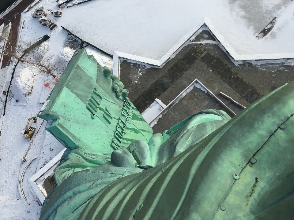 The Statue of Liberty's tablet looking out and down the windows of the crown. It's a sunny day and the grass and trees are green.