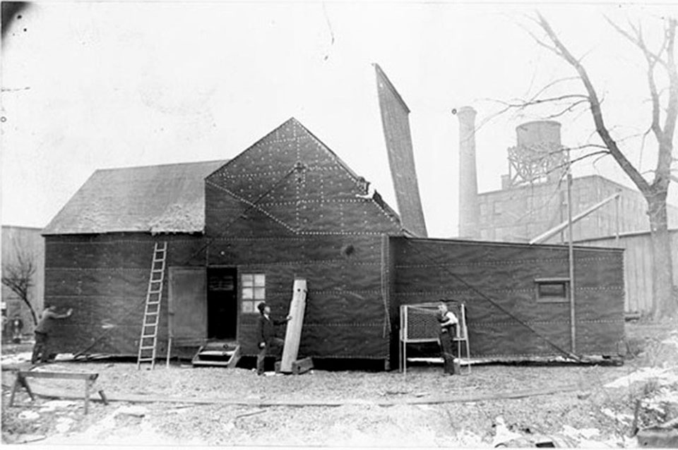 Three workers and a ladder stand at the front of a windowless building that is covered in black tar paper.