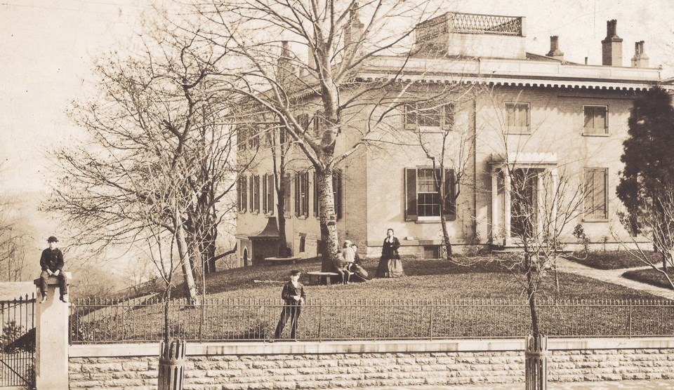 A large two-story home on a hill with several children pictured on the lawn and one child on a fence post