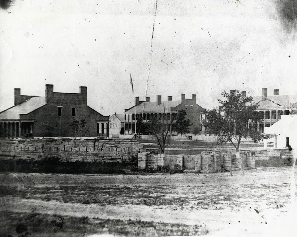 Black and white photo of 3 fort buildings behind a 7 foot stone wall.