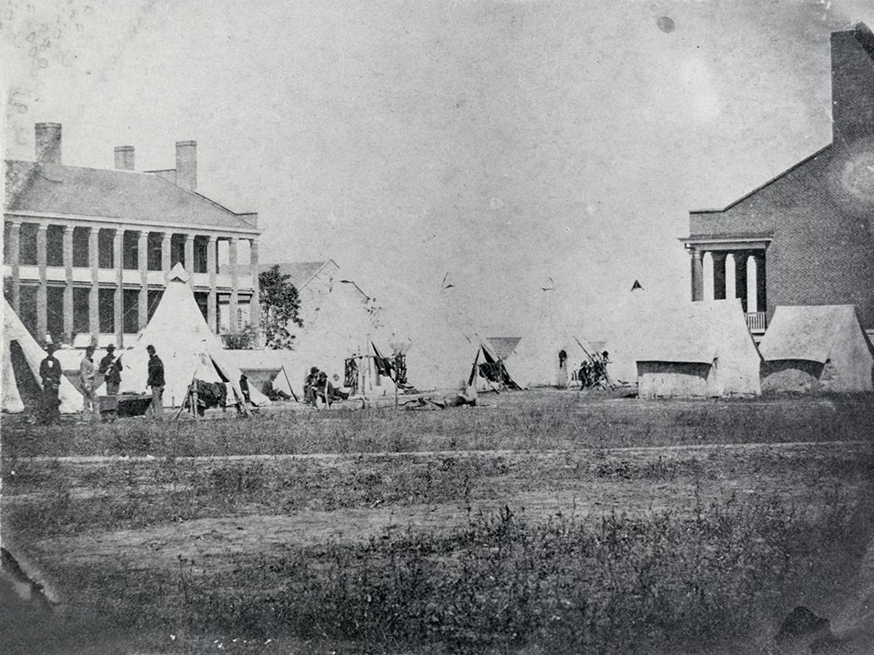 Tents and soldiers camp on the parade ground between fort buildings.