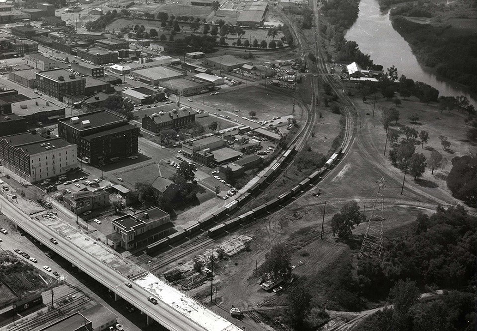 Aerial view with crowded building, roads, and train tracks cutting through park site.