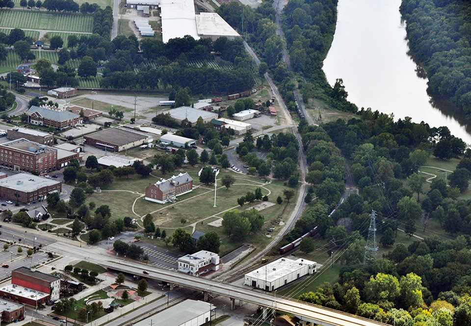 Aerial view with crowded building, roads, and train tracks cutting through park site.