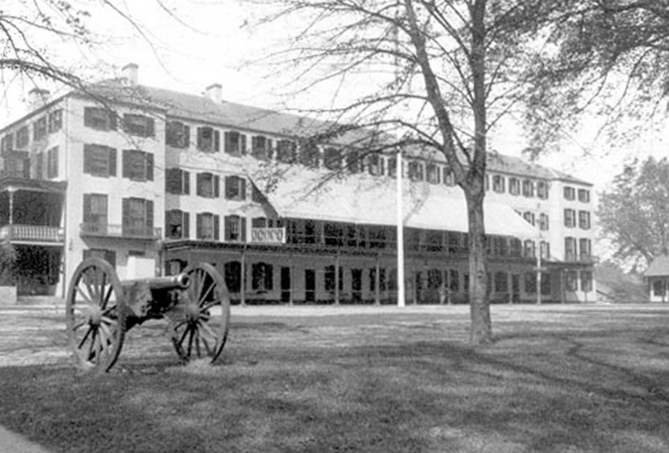 Black and white photo of 3 story rectangular building with cannon in foreground