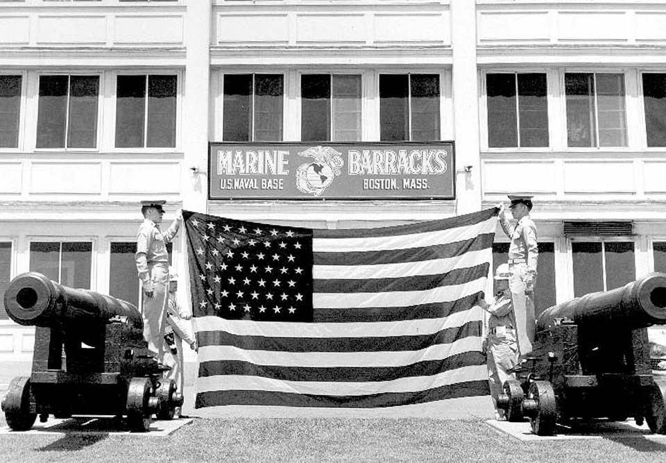 Marine Barracks in backdrop with two marines holding up corners of American Flag, standing on cannon