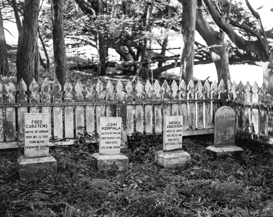 A black and white photo of four grave markers surrounded by a picket fence and trees.