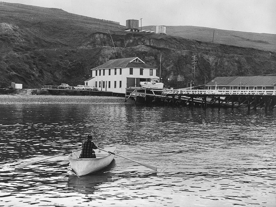 A black and white photo of a man in a plaid, long-sleeved shirt rowing a boat away from a white-sided, two-story boathouse.