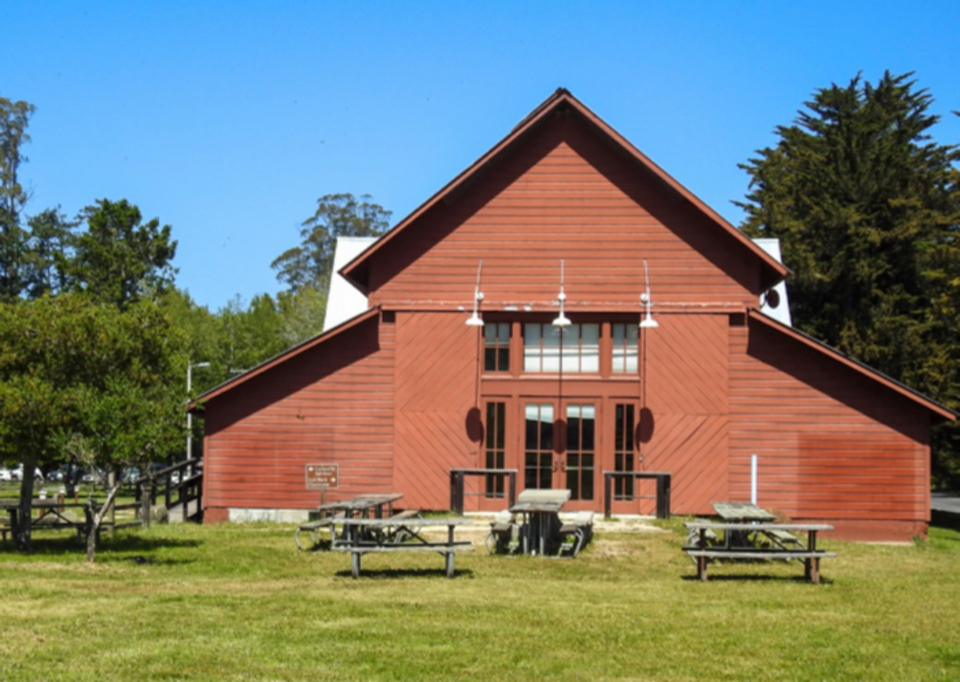 A black and white photo of a barn that is tilted slightly to the right and a broken fence in the foreground.