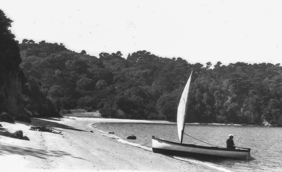 A black and white photo of a man sitting at the stern of a small sailboat as it gently lands on a sandy beach.